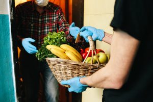 COVID-10 volunteer in a medical mask and gloves passes a basket of food to senior citizen in mask.