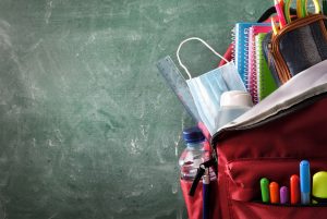 Backpack with school supplies, including mask and sanitizer, with green blackboard in background